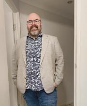 Nick standing in a doorway wearing a beige blazer over a navy wave-pattern shirt